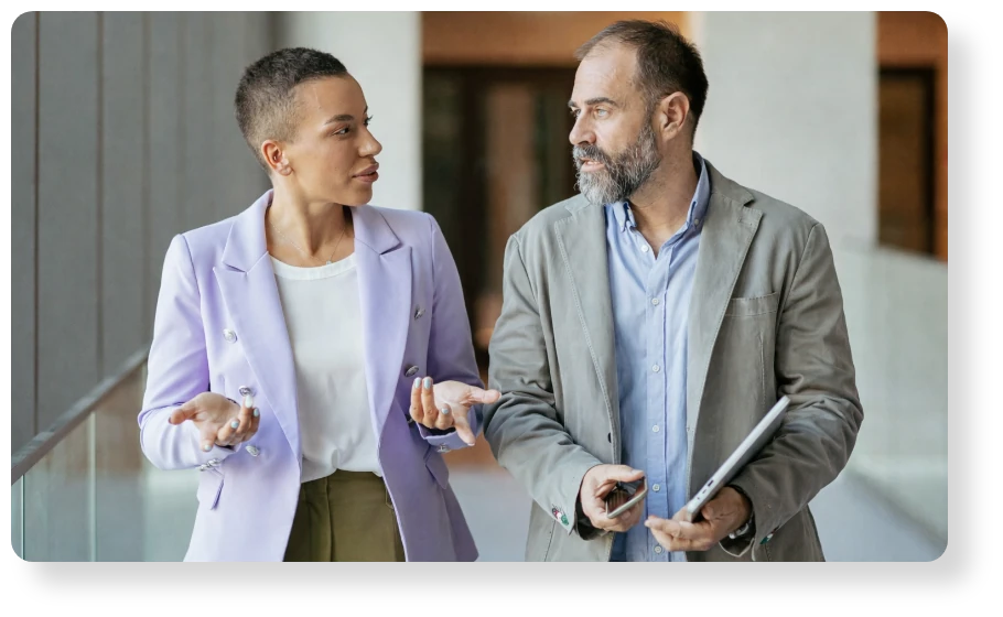 Two business professionals walking indoors and discussing ideas, with one holding a tablet.