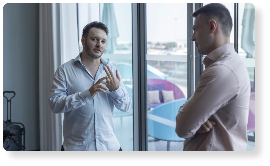 Two business professionals in white shirts having a one-on-one discussion beside large office windows.